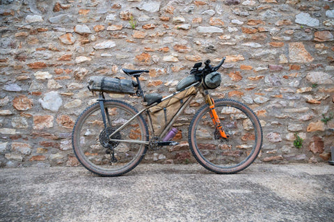 Gravelbike mit Bikepacking-Ausrüstung vor einer Steinmauer, ausgestattet mit Rahmentaschen, Lenkertasche und Satteltasche für lange Touren.