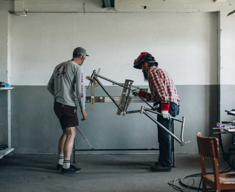 Zwei Männer arbeiten in einer Werkstatt an einem Fahrradrahmen; einer trägt Schweißausrüstung, der andere Freizeitkleidung mit Shorts und Langarmshirt.