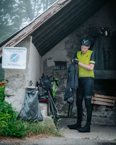 Ein Radfahrer in gelber und schwarzer Radkleidung steht vor einer alten Steinhütte, zieht eine Jacke an und bereitet sich auf eine Tour vor; daneben Fahrräder und Ausrüstung.