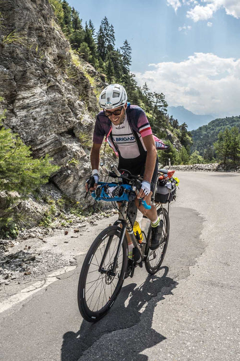 Radfahrer mit Helm und Sonnenbrille fährt ein voll bepacktes Rennrad bergauf auf einer kurvigen Bergstraße bei sonnigem Wetter, umgeben von Felsen und Nadelbäumen.