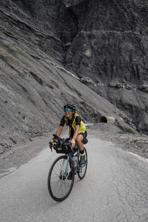 Fahrradfahrerin mit Sonnenbrille und Helm fährt auf einer kurvigen Bergstraße nahe einem Tunnel durch felsige Landschaft.