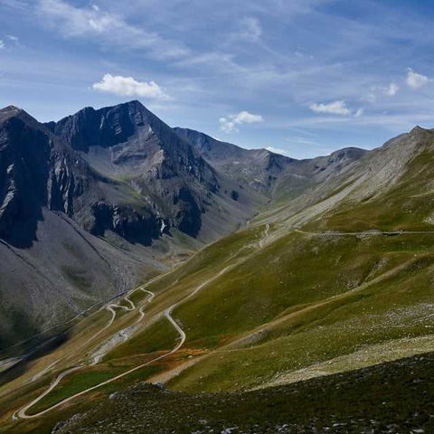 Serpentinenstraße schlängelt sich durch grüne Berghänge in den Alpen unter blauem Himmel mit vereinzelten Wolken. Majestätische Gipfel im Hintergrund.