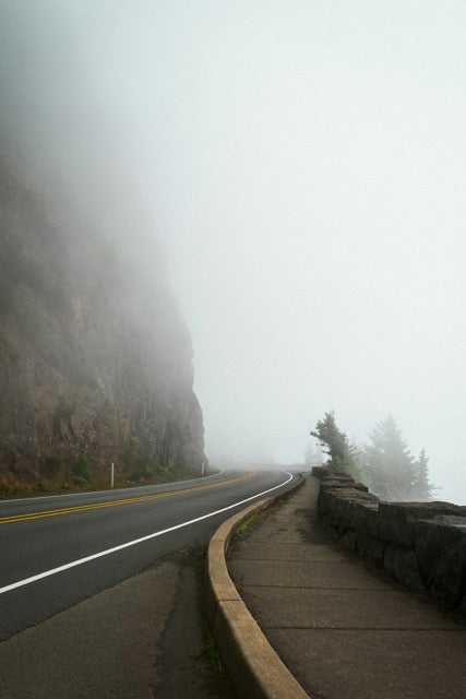 Kurvige Straße an einer Felswand bei dichtem Nebel, mit Gehweg und niedriger Steinmauer auf der rechten Seite, begrenzter Sicht und wenigen Bäumen im Hintergrund.