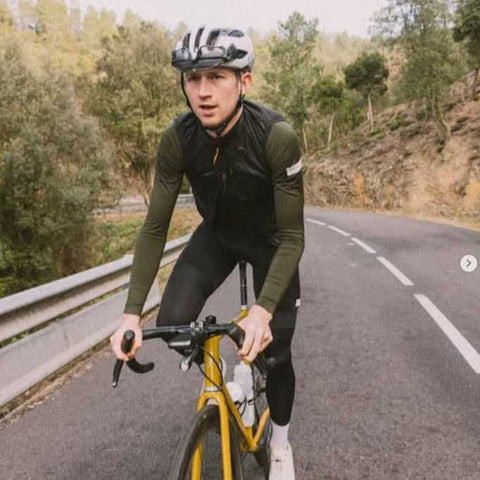 Ein Radfahrer mit Helm und Sportkleidung fährt auf einem gelben Rennrad eine kurvige Straße durch eine bewaldete Hügellandschaft entlang.