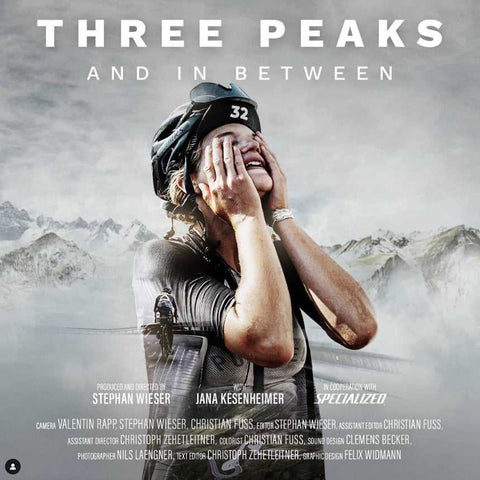 SEO-freundlicher Alt-Text (auf Deutsch):
Glückliche Radfahrerin mit Helm und Startnummer 32 jubelt vor verschneiten Berggipfeln, Filmplakat „Three Peaks and In Between“ mit Namen der Mitwirkenden.