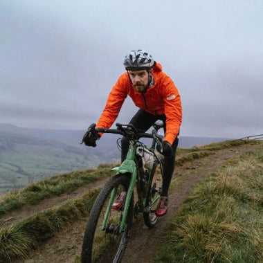 Mann mit roter Jacke fährt mit einem Gravelbike auf einem schmalen, matschigen Bergpfad bei bewölktem Wetter durch hügelige Landschaft.