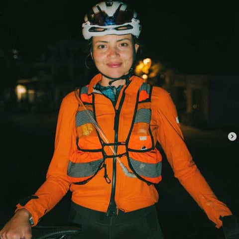 Eine Frau mit Fahrradhelm und orangefarbener reflektierender Weste steht nachts im Freien und lächelt in die Kamera. Sie scheint auf eine Radtour vorbereitet zu sein.