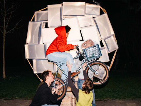 Drei Personen inszenieren eine Szene aus dem Film „E.T. – Der Außerirdische“ mit einem Fahrrad vor einer leuchtenden Mondkulisse; eine Person im roten Hoodie fährt, E.T.-Puppe im Korb.