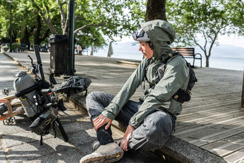 Mann in Outdoor-Bekleidung sitzt auf einer Bordsteinkante an einer Promenade, zieht seine Gamaschen an; neben ihm ein voll bepacktes Reiserad mit Ausrüstung.