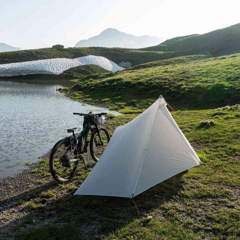 Mensch in Wanderschuhen vor einem weißen Zelt am See, umgeben von Bergen, perfekt für Camping und Outdoor-Aktivitäten in der Natur