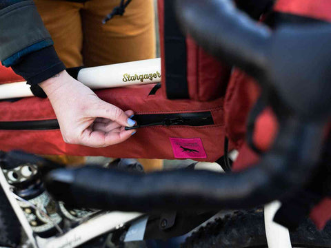 Nahaufnahme einer Hand, die den Reißverschluss der roten Swift Industries / Hold Fast Frame Bag an einem Fahrradrahmen öffnet.