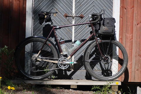 Gravelbike mit Rahmentasche und Trinkflasche lehnt an einer Holztür mit Fischgrätmuster, umgeben von roter Holzfassade und Löwenzahn im Vordergrund.