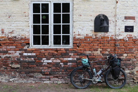 Fahrrad mit Gepäcktaschen lehnt an einer alten Backsteinmauer mit Fenster und Glocke.