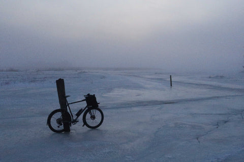 Fahrrad steht angelehnt an einen Pfosten auf zugefrorenem, nebligem See bei Dämmerung oder Morgengrauen.