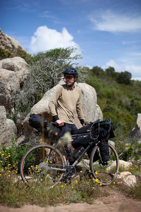 Ein Mann mit Helm und beiger Langarmkleidung steht mit einem voll bepackten Gravelbike auf einem felsigen Pfad inmitten blühender Wildblumen und grüner Hügel.