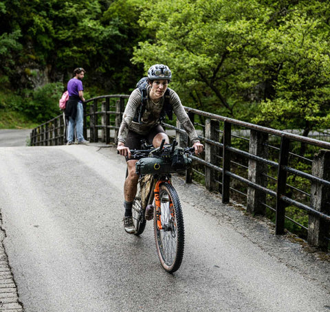 Ein Mountainbiker mit Helm und Ausrüstung fährt über eine Brücke in einer grünen, bewaldeten Landschaft; Kleidung und Fahrrad sind stark verschmutzt vom Gelände.