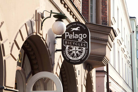 Rundes Geschäftsschild von „Pelago Bicycles“ an einer Hausfassade mit Laterne, zeigt Logo mit Fahrradrad und Schriftzug „Serve the Purpose – Helsinki – Rihimäki – Utilitas – Venustas“.