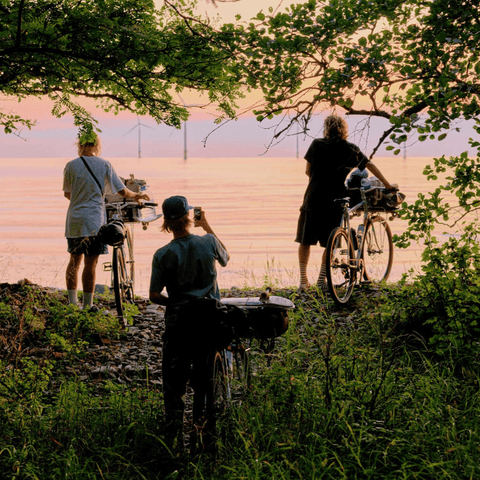 Drei Personen mit Fahrrädern stehen am Ufer eines Sees bei Sonnenuntergang, umgeben von Bäumen und Gras, während eine Person ein Foto macht.