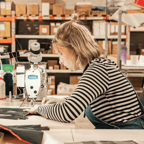 Frau mit blonden Haaren näht konzentriert an einer Industrienähmaschine in einer Werkstatt mit Regalen voller Kartons im Hintergrund.
