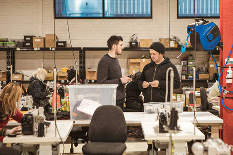 Mehrere Personen arbeiten in einer Werkstatt mit Nähmaschinen und Regalen voller Materialien; zwei Männer stehen im Vordergrund und unterhalten sich.