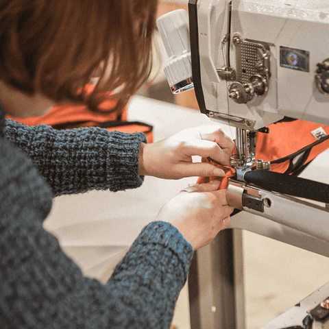 Person näht orangefarbenen Stoff mit einer Industrienähmaschine, Nahaufnahme der Hände beim präzisen Arbeiten in einer Werkstattumgebung.