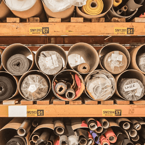 Große Papprollen mit verschiedenen Stoffen und Materialien in einem Lagerregal, beschriftet mit Etiketten und Nummern zur Organisation.