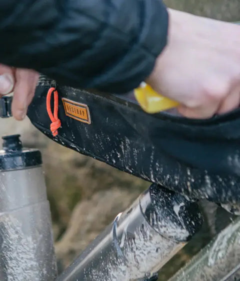 Nahaufnahme einer Hand, die eine schwarze Rahmentasche an einem schlammigen Fahrrad öffnet, mit sichtbarer Trinkflasche und orangefarbenem Reißverschluss-Zugband.