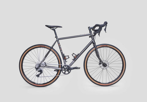 Gravelbike mit schwarzem Stahlrahmen, Rennradlenker, Scheibenbremsen und breiten Reifen mit brauner Seitenwand auf weißem Hintergrund.