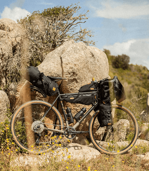 Gravelbike mit umfangreicher Bikepacking-Ausrüstung, darunter Lenkertasche, Rahmentaschen und Gepäcktaschen, abgestellt vor Felsen in einer sonnigen, natürlichen Landschaft.