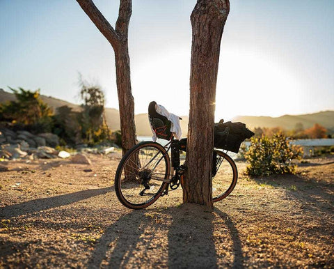Fahrrad lehnt an zwei Bäumen in sonniger Landschaft mit Bergen im Hintergrund, Kleidung über dem Sattel, lange Schatten im warmen Abendlicht.
