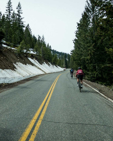 Zwei Radfahrer fahren auf einer kurvigen Bergstraße durch einen Nadelwald mit schmelzendem Schnee am Straßenrand.