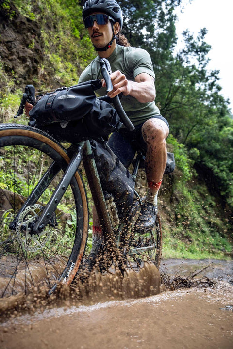 Ein Mountainbiker fährt mit einem Gravelbike durch eine schlammige Pfütze auf einem Waldweg, während Wasser und Schlamm hochspritzen.