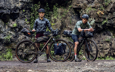 Zwei Bikepacking-Fahrer mit Helmen und Outdoor-Kleidung stehen mit voll bepackten Gravelbikes vor einer Felswand in einer natürlichen Umgebung.