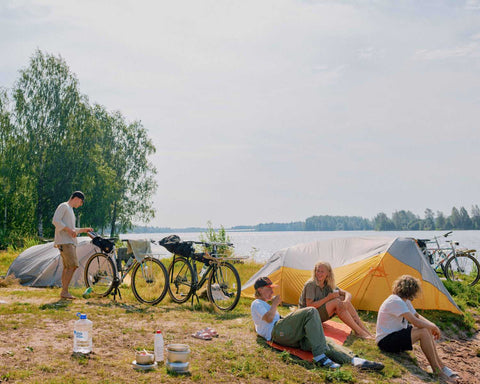 Gruppe junger Menschen beim Zelten am Seeufer mit Fahrrädern, Zelten und Campingausrüstung an einem sonnigen Tag in der Natur.