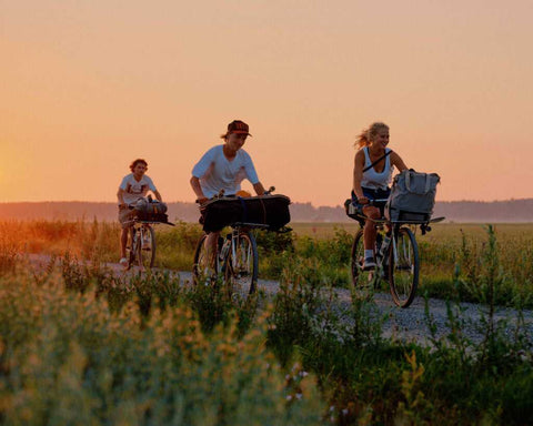 Drei Personen fahren bei Sonnenuntergang mit Fahrrädern auf einem Schotterweg durch eine ländliche Landschaft mit Wiesen und Feldern.