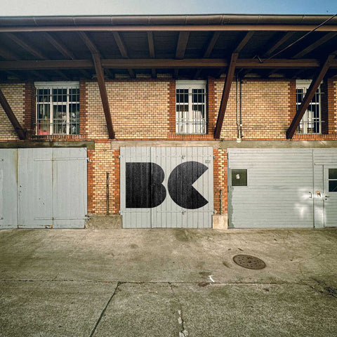 Graffiti mit den Buchstaben „BC“ in schwarzer Farbe auf einem alten Garagentor vor einem Backsteingebäude mit Holzüberdachung und großen Fenstern.