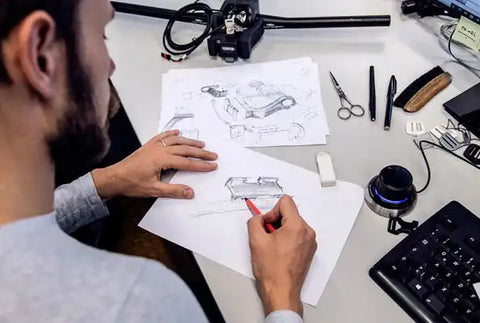 Ein Designer skizziert mit rotem Bleistift ein technisches Produkt auf Papier, umgeben von Zeichnungen, Tastatur, Eingabegerät, Schere und weiteren Büroartikeln auf einem Schreibtisch.