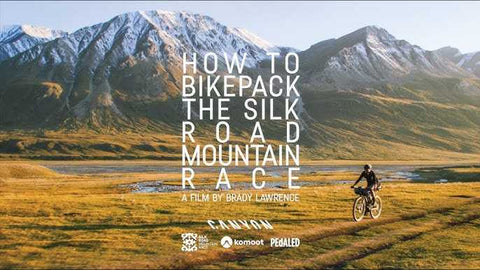 Alt-Text (Deutsch): Radfahrer fährt auf einem Schotterweg vor schneebedeckten Bergen bei Sonnenuntergang; Text: „How to Bikepack the Silk Road Mountain Race – A Film by Brady Lawrence“.