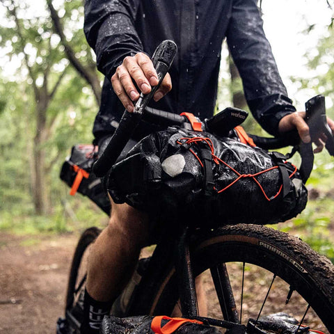 Ortlieb Bike Seat-Pack in Black/Dark Sand montiert am Fahrradlenker bei Regenfahrt im Wald, perfekt für Offroad-Touren.