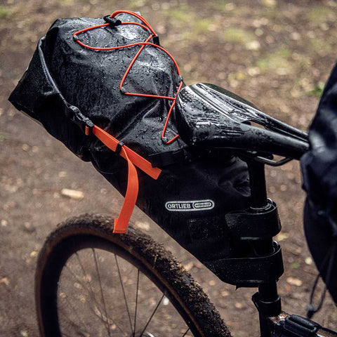 Wasserdichte Ortlieb Satteltasche in Schwarz mit rotem Spanngurt, montiert an einem Fahrrad, bei nassem Wetter im Wald.