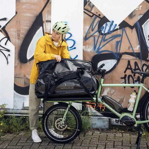 Frau mit Fahrrad in urbaner Umgebung, trägt wasserdichte Ortlieb Duffle RC Tasche auf einem Fahrradträger, ideal für Radfahrer