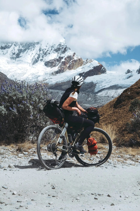 Radfahrer mit Helm und Gepäck auf einem Schotterweg vor schneebedeckten Bergen und blauem Himmel in alpiner Landschaft.