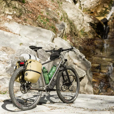 Fahrrad mit gelber Gepäcktasche, auf einer Felsen- und Flusslandschaft, ideal für Outdoor-Abenteuer, aus wasserfestem Material, mit mehreren Wasserflaschen - SKU: 12345