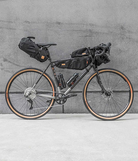 Gravelbike mit kompletter Bikepacking-Ausrüstung, inklusive Rahmentasche, Lenkertasche und Satteltasche, vor grauer Betonwand aufgestellt.