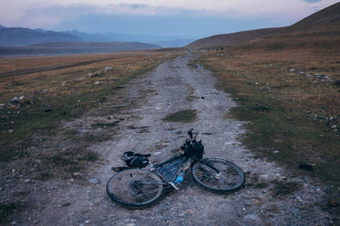 Fahrrad liegt auf einsamer Schotterstraße in bergiger Steppe bei Dämmerung, mit weitem Blick auf entfernte Berge und offenes Gelände.