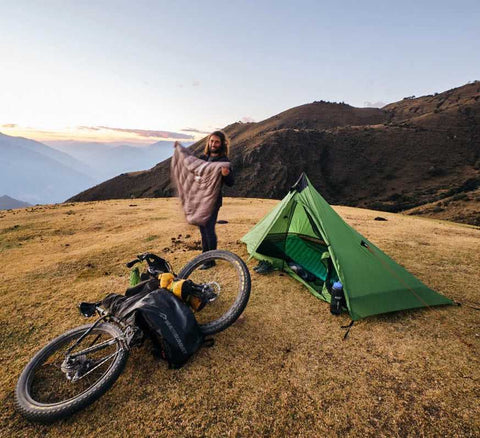 Frau beim Zelten in den Bergen bei Sonnenaufgang, mit Fahrrad und Campingausrüstung auf einer grasbewachsenen Hochebene neben einem grünen Zelt.