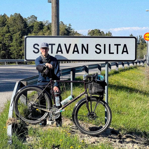 Ein Mann mit Fahrrad steht vor einem Straßenschild mit der Aufschrift „SITAVAN SILTA“ auf einer sonnigen Landstraße mit Wald im Hintergrund.