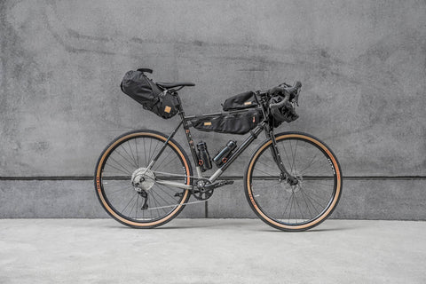 Gravelbike mit kompletter Bikepacking-Ausrüstung, inklusive Rahmentaschen, Lenkertasche und Gepäckträgertasche, vor grauer Betonwand aufgestellt.