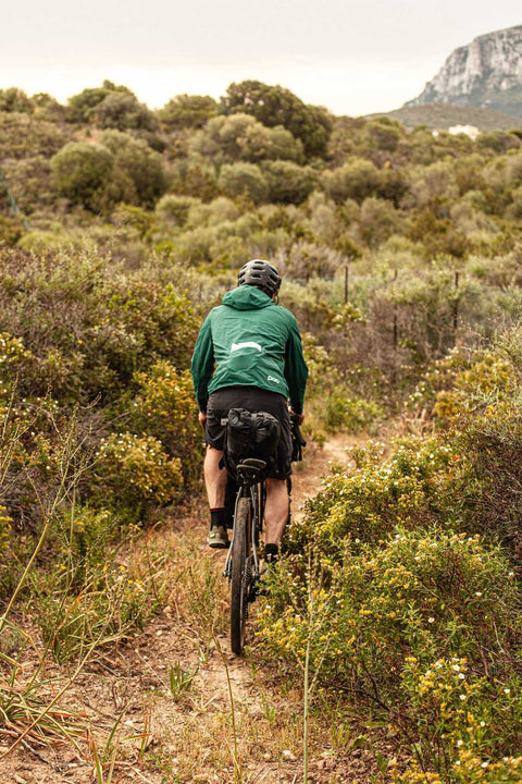 Radfahrer mit Helm und grünem Hoodie fährt auf einem schmalen Naturpfad durch eine mediterrane Landschaft mit Sträuchern und Hügeln im Hintergrund.