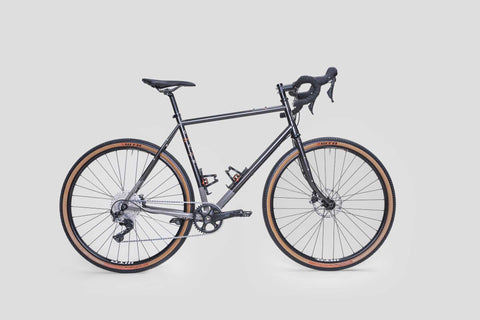 Gravelbike mit schwarzem Stahlrahmen, Rennradlenker, Scheibenbremsen und breiten Reifen mit brauner Seitenwand, geeignet für Straße und Gelände.